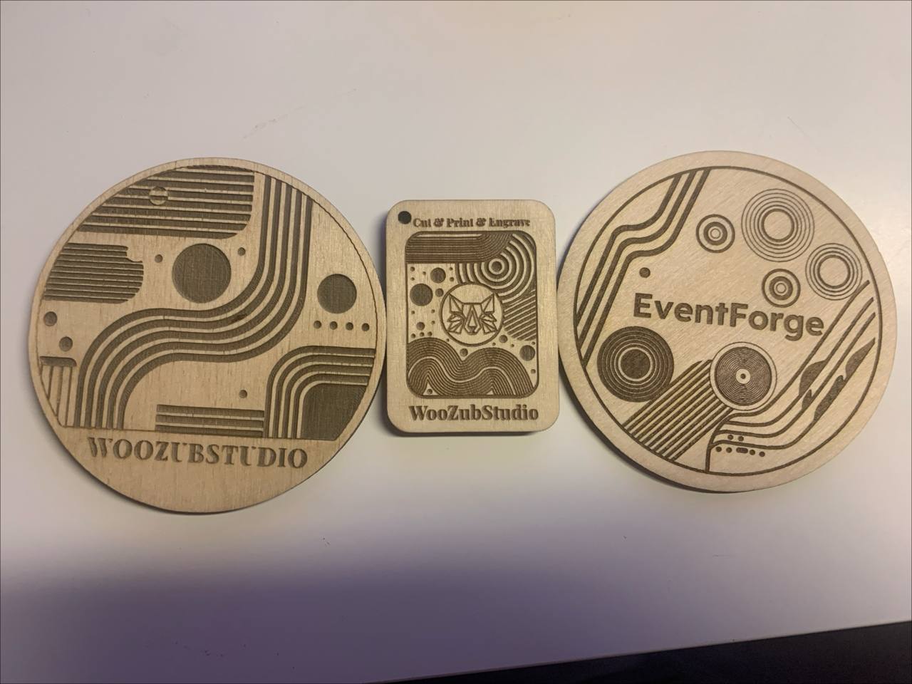 Kolme pyöreää puista lasinalusta, joissa on erilaisia laserkaiverrettuja logoja ja kuvioita.