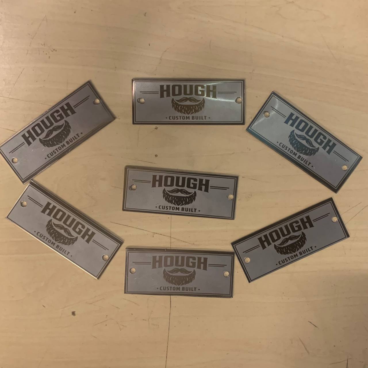 Lähikuva useista valmiista Hough-yrityksen kuitulaserkaiverretuista metallikilvistä, joissa on yrityksen logo ja teksti.